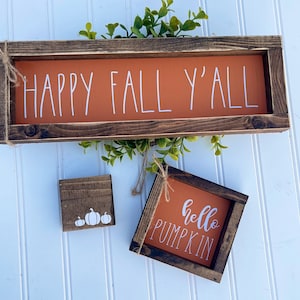May include: Three wooden signs with fall-themed messages. The largest sign is brown with a burnt orange background and says "Happy Fall Y'all". The smaller signs are brown with burnt orange backgrounds and say "hello pumpkin" and feature a white silhouette of three pumpkins.