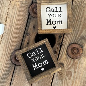 K&ouml;nnte beinhalten: Zwei kleine, quadratische Holzschilder mit dem Text "Call Your Mom" in schwarzer Schrift. Ein Schild hat einen wei&szlig;en Hintergrund, das andere einen schwarzen Hintergrund. Beide Schilder haben ein kleines Herzsymbol und sind in einem rustikalen, braunen Holzrahmen gefasst.