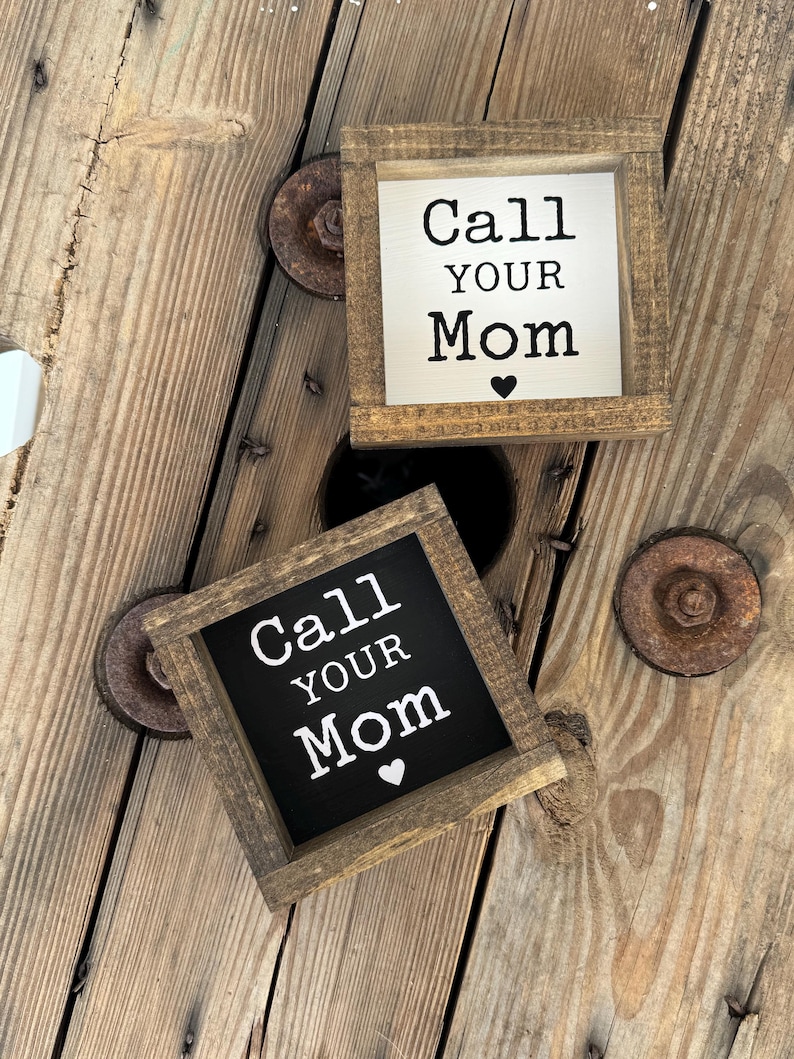 K&ouml;nnte beinhalten: Zwei kleine, quadratische Holzschilder mit dem Text "Call Your Mom" in schwarzer Schrift. Ein Schild hat einen wei&szlig;en Hintergrund, das andere einen schwarzen Hintergrund. Beide Schilder haben ein kleines Herzsymbol und sind in einem rustikalen, braunen Holzrahmen gefasst.