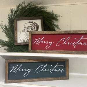 May include: Two wooden signs with the text "Merry Christmas" in white script. The top sign has a red background and the bottom sign has a dark blue background. The signs are on a white shelf with a green garland behind them.