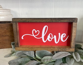 May include: A red wooden sign with a white heart and the word "love" in a cursive font. The sign is framed in a dark brown wood.