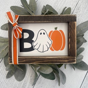 May include: A wooden sign with a black and white "BOO" with a ghost and an orange pumpkin. The sign is framed in dark wood and has a white background.