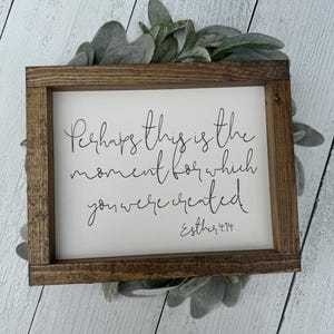 May include: A rectangular wooden sign with the quote "Perhaps this is the moment for which you were created" in black script. The sign is framed in dark brown wood and surrounded by green and grey foliage.