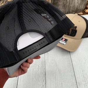 Puede incluir: Gorra de camionero gris y negra con la parte trasera de malla negra y cierre a presi&oacute;n. La gorra se sostiene, revelando la parte inferior. Otra gorra con la parte delantera marr&oacute;n y la parte trasera de malla negra est&aacute; en el fondo.