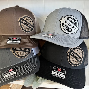 May include: A stack of six trucker hats in various colors, including brown, gray, and black. Each hat features a mesh back and a front patch that reads "OSHA CERTIFIED VIOLATOR". The hats are adjustable and have a Richardson 112 tag.