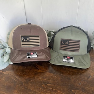 May include: Two baseball caps with a brown and green mesh back. The front of the caps are brown and green with a white and red American flag with a brown deer antler design. The caps have a black label with the text "Richardson 112" on them.