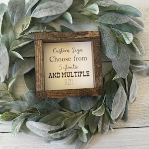 May include: A small, square wooden sign with the text "Custom Sign Choose from 5 fonts AND MULTIPLE SIZES" is centered on a green and gray leafy wreath. The sign has a dark brown frame.
