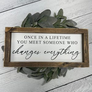 May include: A rectangular wooden sign with a white background and a dark brown frame. The sign reads "ONCE IN A LIFETIME YOU MEET SOMEONE WHO changes everything" in black script. The sign is decorated with green leaves.