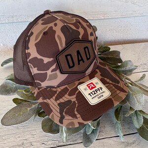 May include: A brown camouflage baseball cap with a mesh back. The cap features a black patch with the word "DAD" and a white tag with the text "112PFP PRINTED 5 PANEL OSFM".