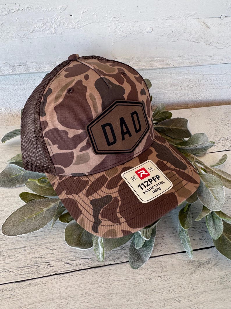 May include: A brown camouflage baseball cap with a mesh back. The cap features a black patch with the word "DAD" and a white tag with the text "112PFP PRINTED 5 PANEL OSFM".