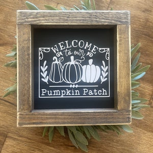 May include: A rustic wooden framed sign with a black chalkboard insert. The sign says "Welcome to our Pumpkin Patch" with a white silhouette of three pumpkins and leaves.