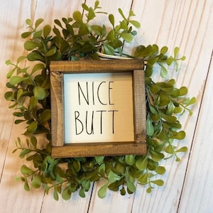 May include: A small wooden sign with a white background and black text that reads "NICE BUTT". The sign is framed in a dark brown wood frame and is surrounded by green foliage.