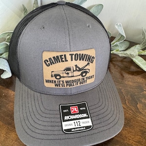 May include: Gray trucker hat with a black mesh back. The front features a tan leather patch with the words "CAMEL TOWING" and a graphic of a tow truck. Text below reads "WHEN IT'S WEDGED IN TIGHT WE'LL PULL IT OUT!" A Richardson 112 Trucker tag is visible.