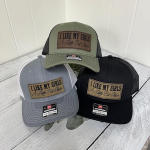 May include: Three trucker hats in olive green, gray, and black. Each hat has a mesh back and a rectangular patch with the text "I LIKE MY GIRLS A Little Bit Older". The hats are adjustable and have a Richardson 112 tag.