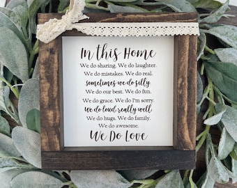 Op de afbeelding: Een houten bord met een witte achtergrond en zwarte tekst die luidt "In this home we do sharing, we do laughter, we do mistakes, we do real, sometimes we do silly, we do our best, we do fun, we do grace, we do I'm sorry, we do loud really well, we do hugs, we do family, we do awesome, we do love". Het bord is omlijst met een donkerbruin houten frame en heeft een witte kantversiering.