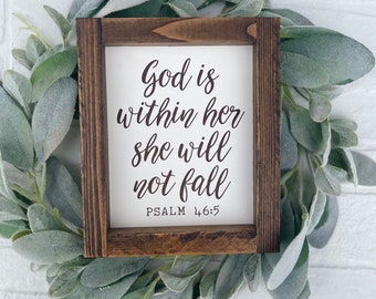 May include: A wooden sign with a white background and brown text. The sign reads "God is within her she will not fall Psalm 46:5". The sign is surrounded by a wreath of green leaves.