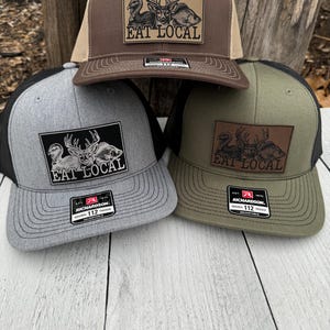 May include: Three trucker caps in brown, grey, and olive green. Each cap has a rectangular patch with a deer, fish, and bird illustration, and the text "EAT LOCAL". The caps have a mesh back and a curved peak.