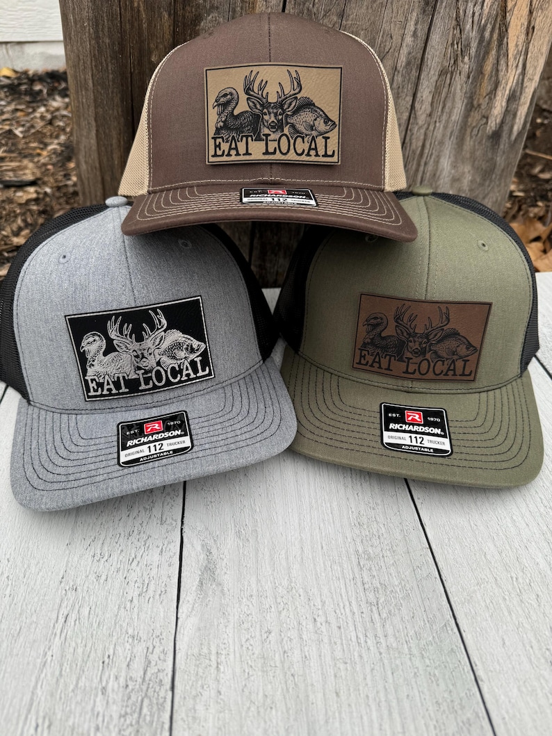 Puede incluir: Tres gorras de camionero en marr&oacute;n, gris y verde oliva. Cada gorra presenta un parche rectangular con una ilustraci&oacute;n de ciervo, pez y p&aacute;jaro, y el texto "EAT LOCAL". Las gorras tienen una parte trasera de malla y una visera curva.