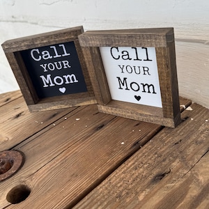 K&ouml;nnte beinhalten: Zwei kleine, quadratische Holzschilder mit dem Text "Call Your Mom" in wei&szlig;en Buchstaben. Ein Schild hat einen schwarzen Hintergrund, das andere einen wei&szlig;en. Beide haben ein kleines Herzsymbol und sind in einem dunkelbraunen Holzrahmen gefasst.