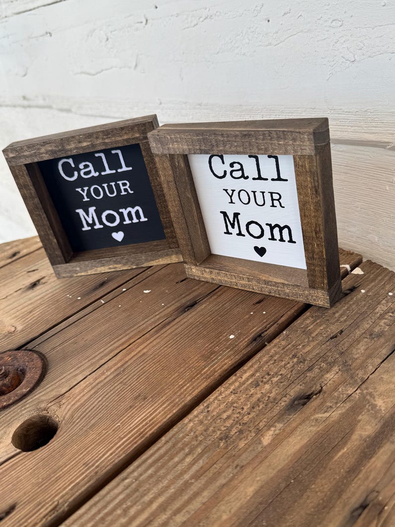 K&ouml;nnte beinhalten: Zwei kleine, quadratische Holzschilder mit dem Text "Call Your Mom" in wei&szlig;en Buchstaben. Ein Schild hat einen schwarzen Hintergrund, das andere einen wei&szlig;en. Beide haben ein kleines Herzsymbol und sind in einem dunkelbraunen Holzrahmen gefasst.