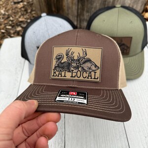 Puede incluir: Gorra de camionero marr&oacute;n y beige con un parche que muestra un pavo, un ciervo y un pez, con el texto "EAT LOCAL". La gorra tiene una visera curva y una parte trasera de malla. La gorra de camionero Richardson 112 es ajustable.