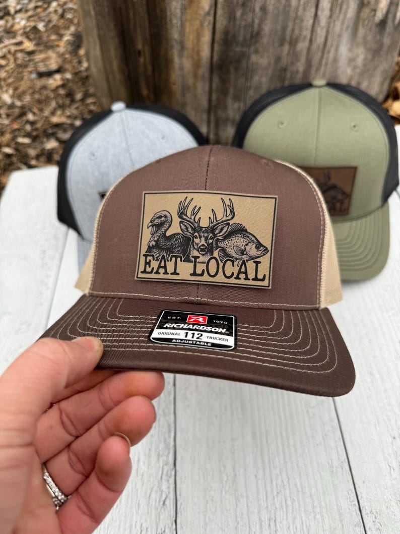 Puede incluir: Gorra de camionero marr&oacute;n y beige con un parche que muestra un pavo, un ciervo y un pez, con el texto "EAT LOCAL". La gorra tiene una visera curva y una parte trasera de malla. La gorra de camionero Richardson 112 es ajustable.