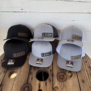 Peut inclure: Plusieurs casquettes de baseball noires et grises avec des dos en filet. Chaque casquette porte un écusson rectangulaire avec les mots "LEGEND" ou "LEGACY". Les casquettes sont exposées sur une bobine en bois.