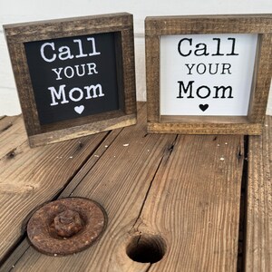 K&ouml;nnte beinhalten: Zwei kleine, quadratische Holzschilder mit dem Text "Call Your Mom" in wei&szlig;er Schrift. Ein Schild hat einen schwarzen Hintergrund, das andere einen wei&szlig;en Hintergrund. Beide haben ein kleines Herzsymbol. Die Schilder stehen auf einer Holzoberfl&auml;che.