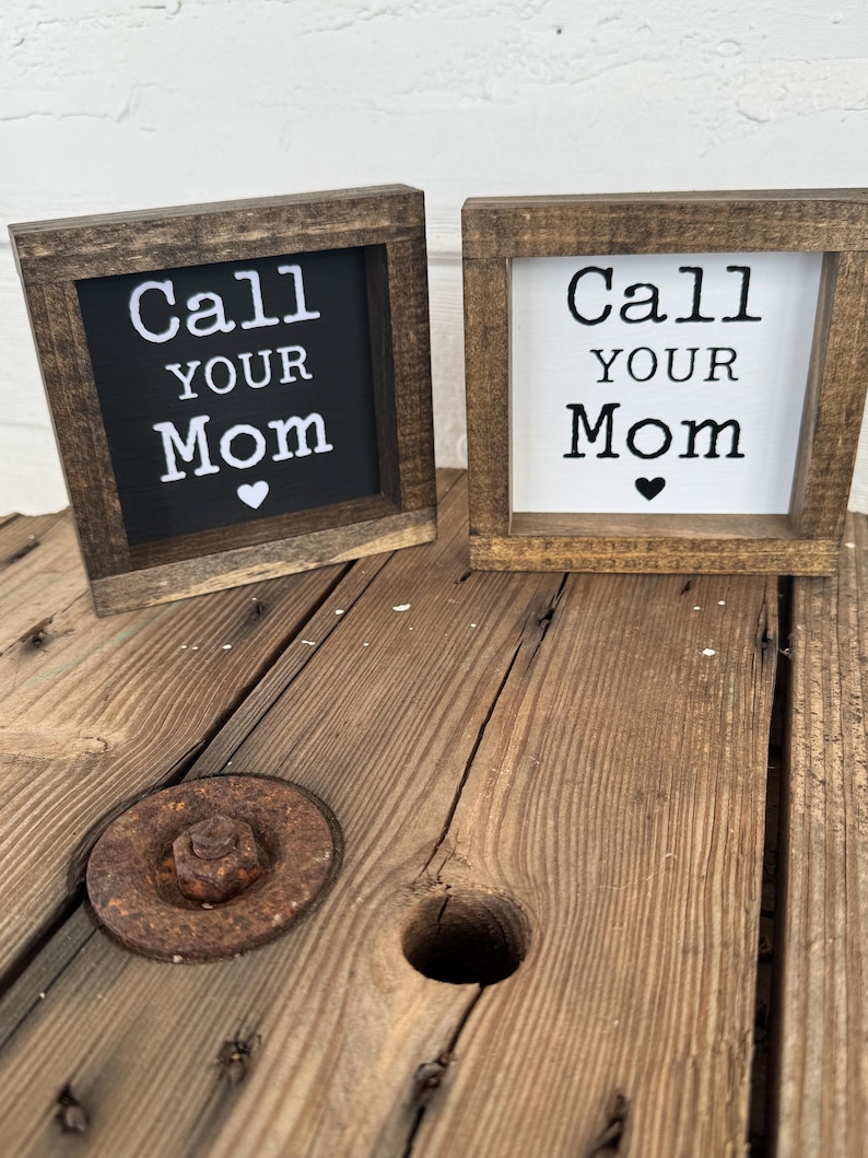 K&ouml;nnte beinhalten: Zwei kleine, quadratische Holzschilder mit dem Text "Call Your Mom" in wei&szlig;er Schrift. Ein Schild hat einen schwarzen Hintergrund, das andere einen wei&szlig;en Hintergrund. Beide haben ein kleines Herzsymbol. Die Schilder stehen auf einer Holzoberfl&auml;che.