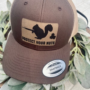 May include: A brown and tan trucker hat with a brown leather patch that says "Protect Your Nuts" and a silhouette of a squirrel holding a gun. The hat has a mesh back and a YUPOONG logo.