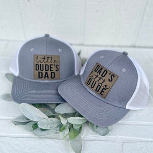 Peut inclure: Deux casquettes de camionneur grises et blanches avec des patchs marrons. La plus grande casquette dit "Dad's little dude" et la plus petite dit "little dude's dad".