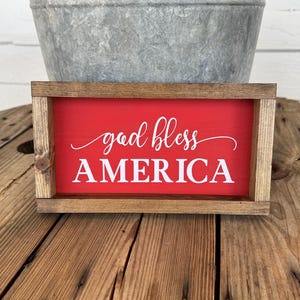 Puede incluir: Un letrero rectangular de madera con fondo rojo y las palabras "god bless AMERICA" en escritura blanca. El letrero tiene un marco de madera natural y se muestra sobre una superficie de madera.