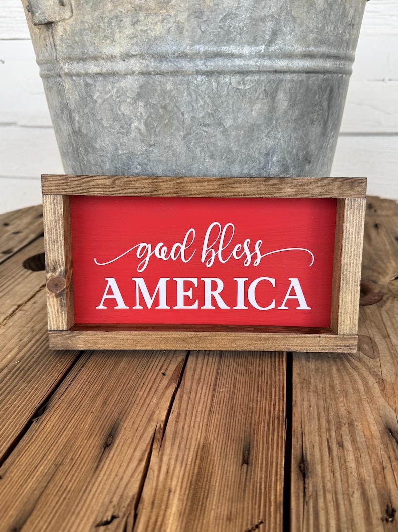 Puede incluir: Un letrero rectangular de madera con fondo rojo y las palabras "god bless AMERICA" en escritura blanca. El letrero tiene un marco de madera natural y se muestra sobre una superficie de madera.