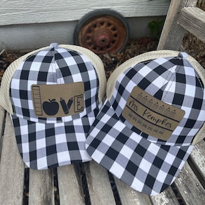 May include: Two black and white checkered trucker hats with tan mesh backs. One hat features a brown leather patch with the word "LOVE" and school-themed graphics. The other hat has a patch with the name "Ms. Kempkes" and pencil graphics.