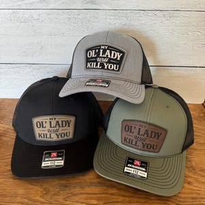 May include: Three trucker caps in black, grey, and olive green, each with a mesh back and a curved peak. The caps feature a patch with the text "MY OL' LADY WILL KILL YOU."