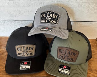 May include: Three trucker caps in black, grey, and olive green, each with a mesh back and a curved peak. The caps feature a patch with the text "MY OL' LADY WILL KILL YOU."