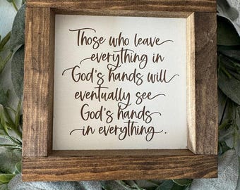May include: A square wooden sign with a brown frame. The sign has the text "Those who leave everything in God's hands will eventually see God's hands in everything" in brown script. The sign is surrounded by green and gray foliage.
