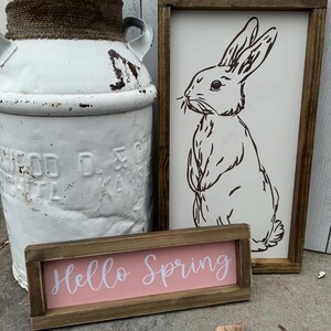 May include: A rustic white milk can with a rope accent, a wooden sign with a bunny illustration, and a small sign that reads "Hello Spring" in white script on a pink background. The signs have brown wooden frames.