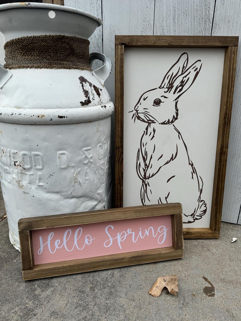 May include: A rustic white milk can with a rope accent, a wooden sign with a bunny illustration, and a small sign that reads "Hello Spring" in white script on a pink background. The signs have brown wooden frames.