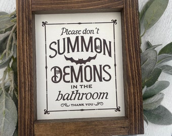 May include: A wooden framed sign with a white background and black text that reads "Please don't summon demons in the bathroom. Thank you."