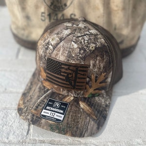 Peut inclure: Un chapeau de camionneur camouflage brun et beige avec un patch en cuir brun représentant un drapeau américain et une silhouette de lineman sur un poteau électrique. Le chapeau a un dos en maille noire et une sangle réglable noire avec le texte "EST. 1970 RICHARDSON. ORIGINAL 112 TRUCKER ADJUSTABLE" imprimé dessus.