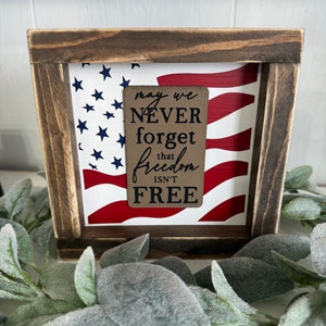 May include: A wooden framed sign with an American flag design and a brown tag with the text "may we NEVER forget that freedom ISN'T FREE".