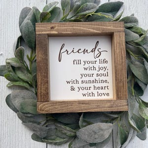 May include: A square wooden sign with a dark brown frame and a white center panel. The text in dark brown script and print reads: friends fill your life with joy, your soul with sunshine, & your heart with love. The sign is surrounded by green leaves.