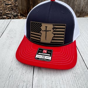 May include: A navy blue and white trucker hat with a red brim. The hat features a leather patch with an American flag design and a cross. The hat has the text "RICHARDSON ORIGINAL 112 TRUCKER ADJUSTABLE".