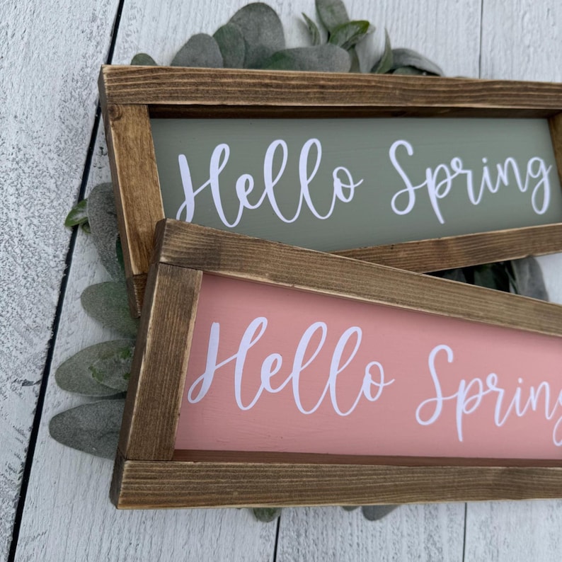 May include: Two rectangular wooden signs with the words "Hello Spring" in white script. One sign has a pink background, the other a green background. The signs are framed in brown wood and rest on a bed of green leaves.