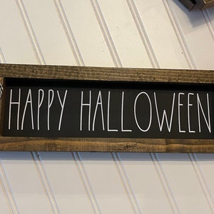 May include: A wooden sign with a black background and white lettering that reads "Happy Halloween". The sign is framed in a dark brown wood and has a black and white checkered ribbon attached to the top.