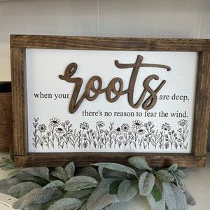 May include: A wooden sign with a brown frame and a white background. The sign reads "When your roots are deep, there's no reason to fear the wind." The word "roots" is in a large, decorative font and is made of wood. There are small, black drawings of flowers and leaves at the bottom of the sign.