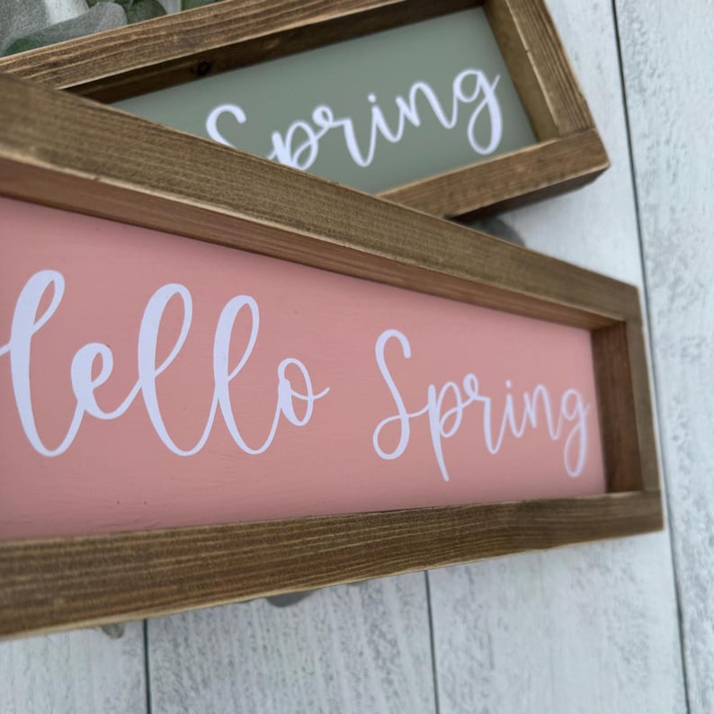May include: Two rectangular wooden signs with the words "Hello Spring" and "Spring" in white script. The signs have a light pink and light green background, respectively, and are set in a rustic brown wooden frame.