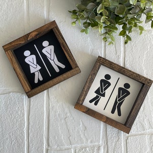 May include: Two wooden signs with black and white stick figures indicating the directions to the men's and women's restrooms. The signs are framed in dark brown wood.