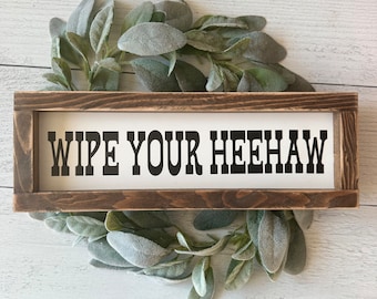 May include: A rustic wooden sign with the text "Wipe Your Heehaw" in black lettering. The sign is surrounded by a wreath of green and gray foliage.